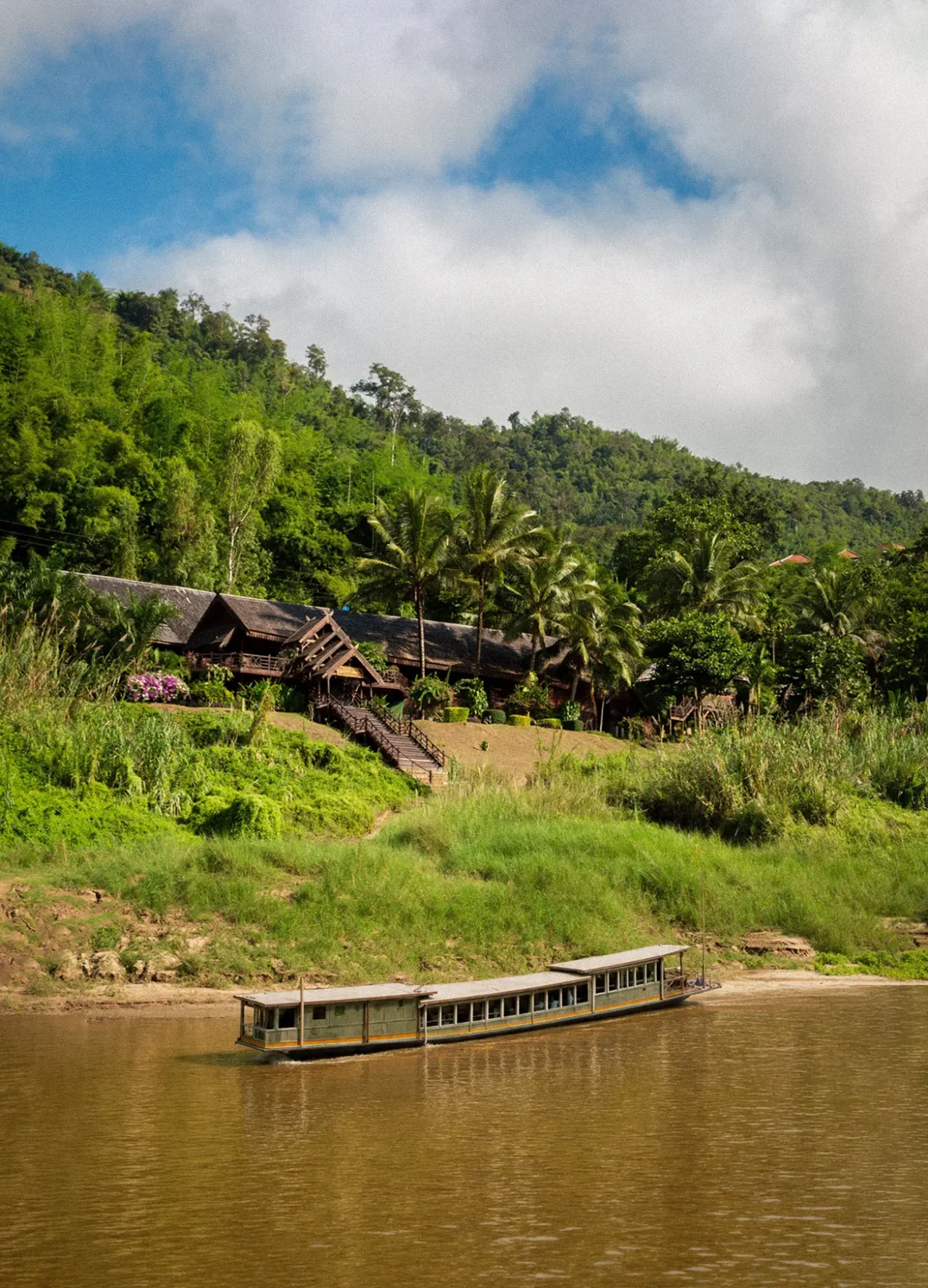 A Mekong Cruises boat on the river below Luang Say Lodge, its teak buildings on a forested hillside