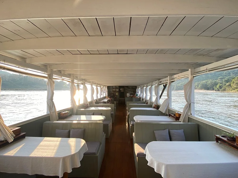The boat’s saloon in the late-afternoon sun: tables and cushioned benches along open windows on the Mekong